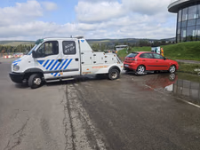 Tractare autoturism mic — un hatchback roșu (Seat Ibiza), defect, ridicat pe platforma cu cârlig a mașinii de tractare, lângă o clădire modernă cu fațadă vitrată. Tractare autoturism mic — un hatchback roșu (Seat Ibiza), defect, ridicat pe platforma cu cârlig a mașinii de tractare, lângă o clădire modernă cu fațadă vitrată.
