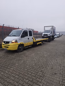Zi cu ceață, curte industrială. Un camion tip Ford Transit cu cutie de marfă este încărcat integral pe platforma galbenă a mașinii de tractare, lângă alte autoutilitare aliniate. Zi cu ceață, curte industrială. Un camion tip Ford Transit cu cutie de marfă este încărcat integral pe platforma galbenă a mașinii de tractare, lângă alte autoutilitare aliniate.